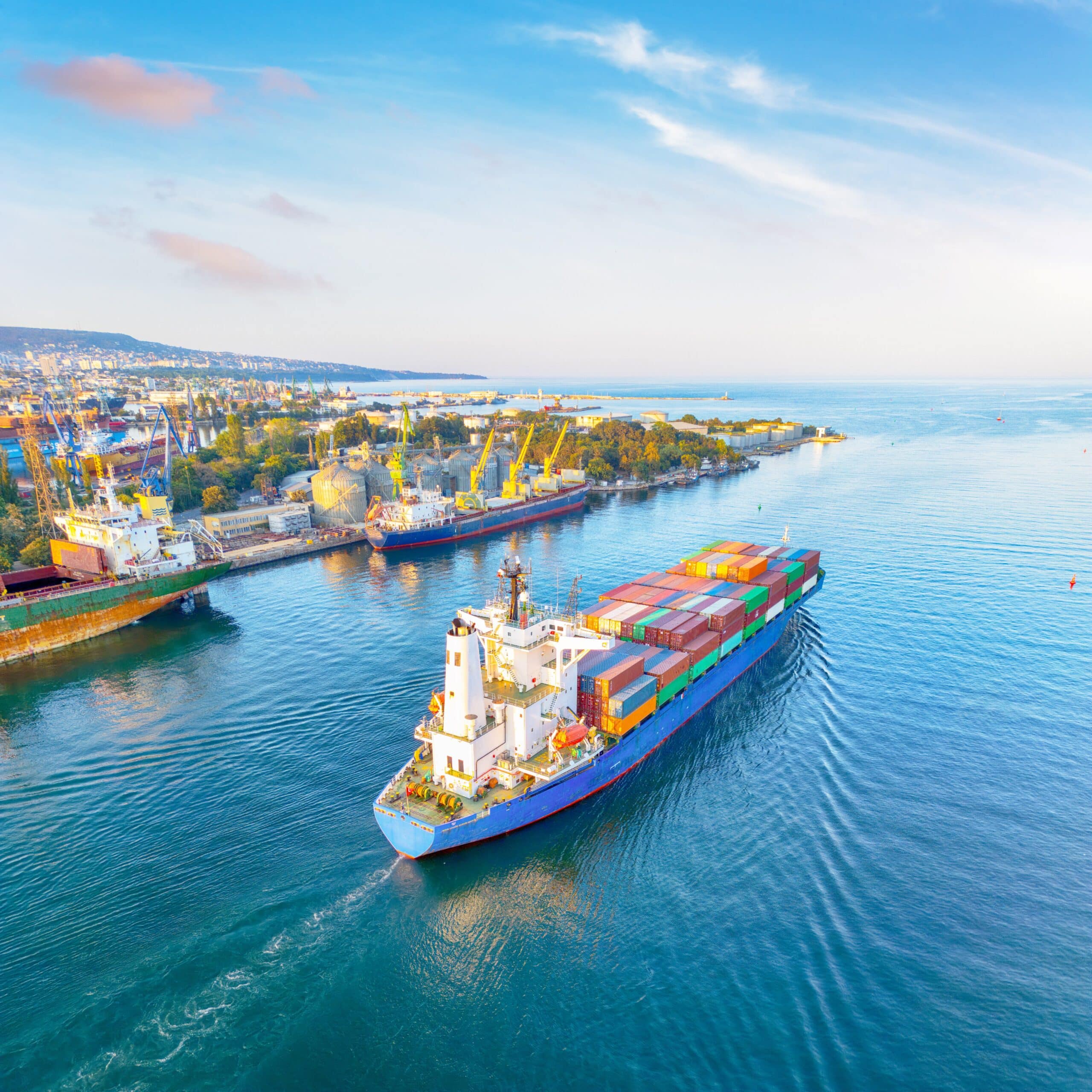 Ship with lots of colorful cargo on it moving through the water alongside a port with other cargo ships